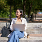 Portrait of korean woman sitting with laptop in park on bench, working outdoors, student doing homework, drinking takeaway coffee.