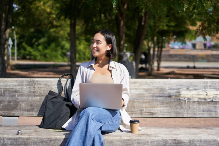 Portrait of korean woman sitting with laptop in park on bench, working outdoors, student doing homework, drinking takeaway coffee.